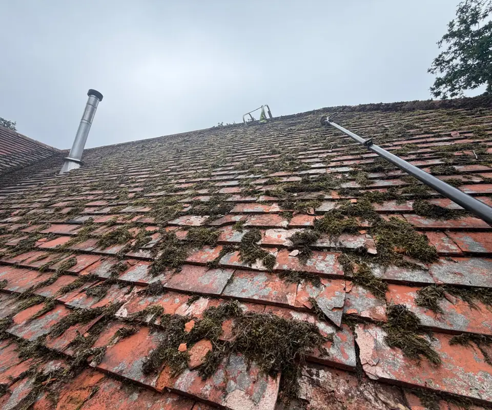 Looking up a mossy red tile roof towards a metal flue, with a long pole up the slope.